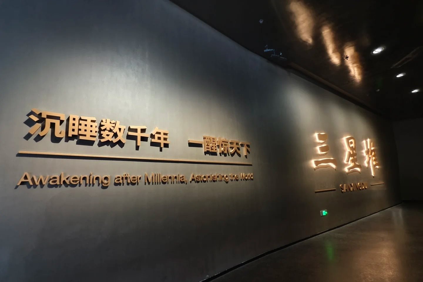 A dark grey wall inside the museum carrying raised gold characters reading 沉睡数千年 一醒惊天下, with the English line 'Awakening after Millennia, Astonishing the World' beneath it, and the illuminated museum name 三星堆 SANXINGDUI further along the wall.
