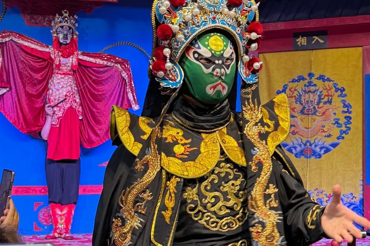 Sichuan opera performer in a green painted mask, pearl headdress and gold-embroidered black robe standing beside the guest rail