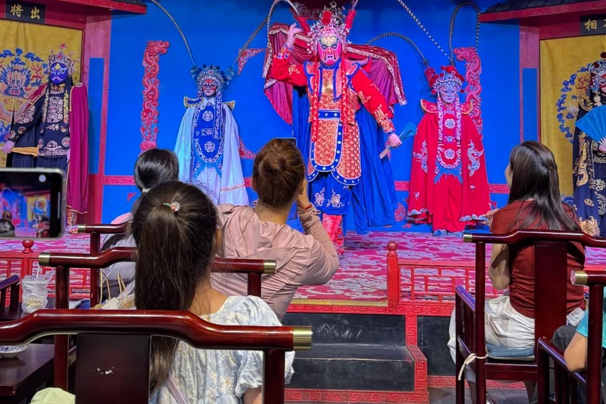 Audience in wooden armchairs with tea tables watching three costumed Sichuan opera performers on a small Chengdu stage