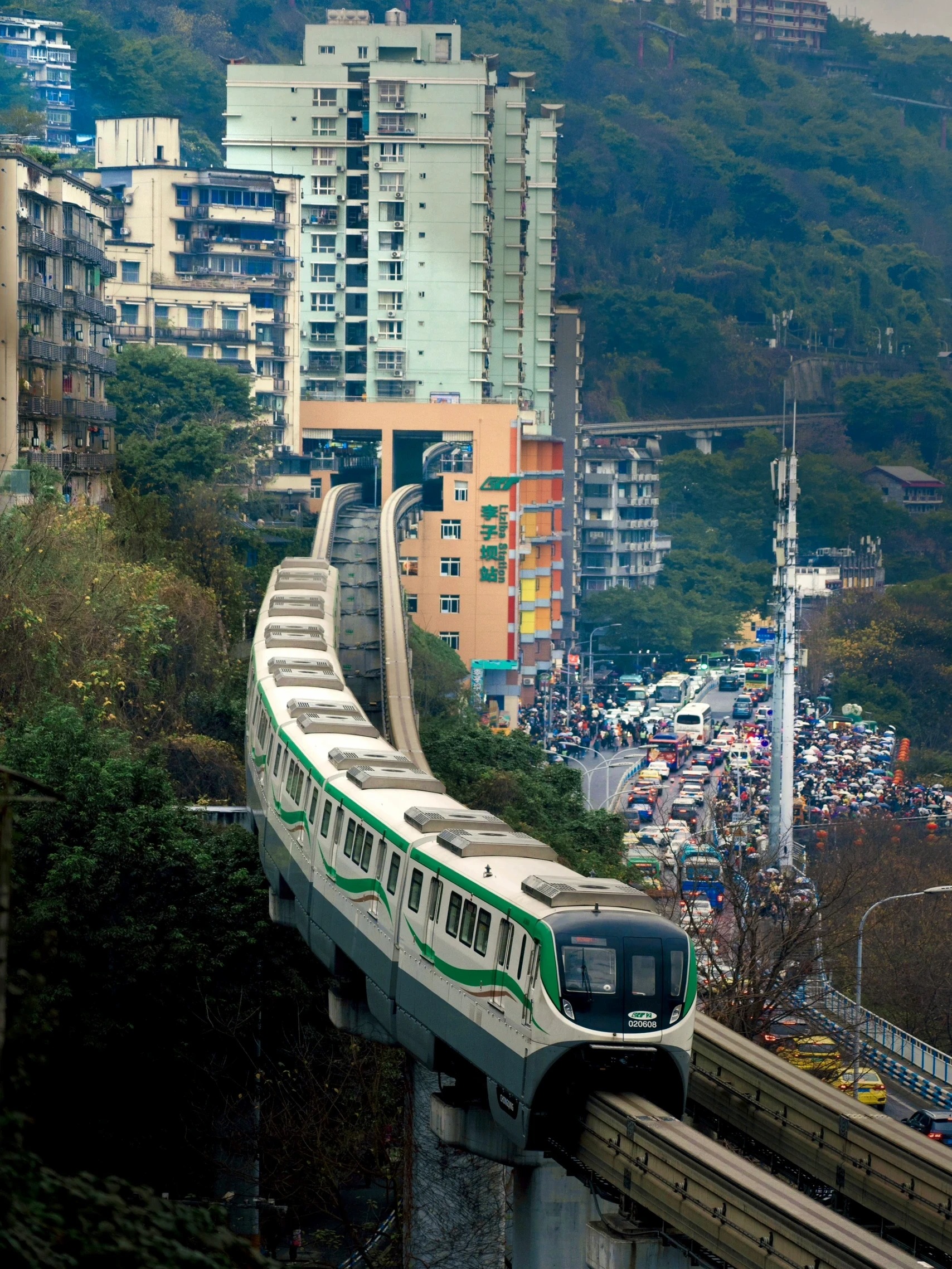 The Liziba monorail curving out of the hillside with the station sign on an orange building, and a dense crowd of visitors under umbrellas filling the street beside the viewing area.