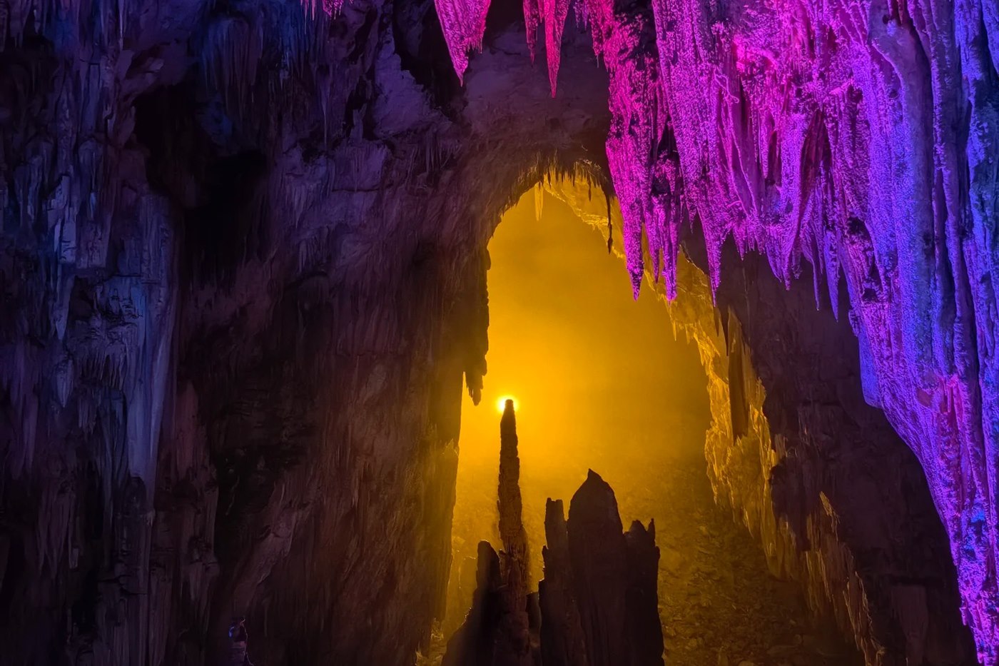Inside Furong Cave — a tall natural archway lit gold from behind, with stalactites overhead lit magenta and violet and a single stalagmite silhouetted in the opening.