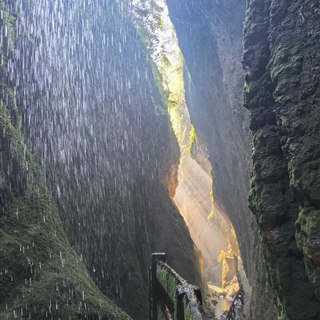 A very narrow Wulong fissure with water falling in a curtain across the gap, sunlight striking the far wall and a walkway railing below.