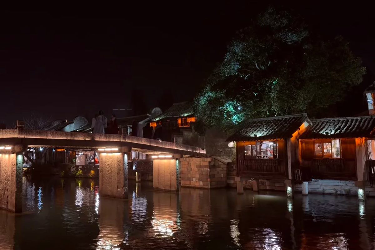 A lit stone beam bridge across a Wuzhen canal after dark, its piers uplit and mirrored in still water, with timber water-houses along the right bank showing warm lamplit windows and a handful of silhouetted figures crossing the bridge.