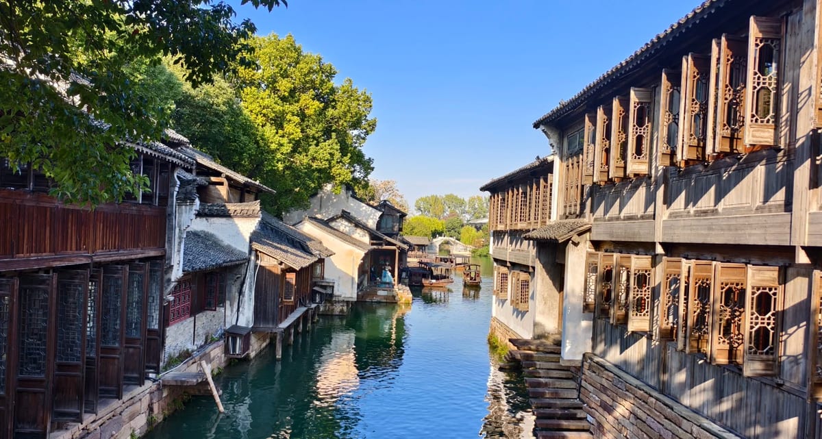 A canal in Wuzhen under a blue sky, lined on both banks with two-storey dark-timber stilt houses built out over the water, with wooden boats moored below and a stone arch bridge in the distance.