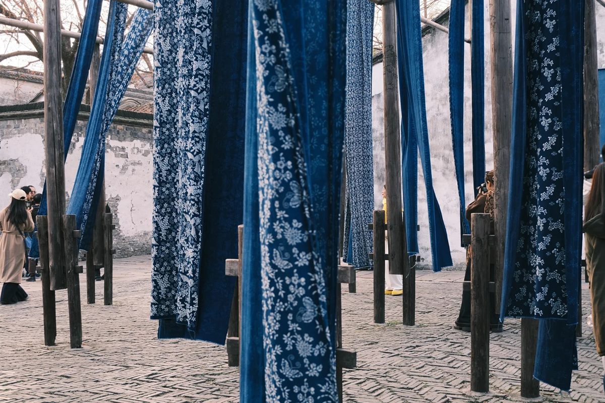 Rows of tall weathered timber poles in a Wuzhen dye works, hung with long lengths of blue-and-white indigo-printed cloth drying above a brick-paved yard, with the whitewashed courtyard wall behind.