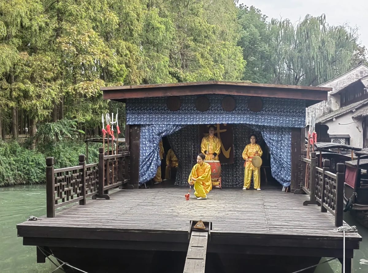 A martial-arts performance on a moored stage boat in Wuzhen: four performers in yellow silk in front of an indigo-print backdrop bearing a large gold character for martial, with a drum, a gong and a rack of weapons.