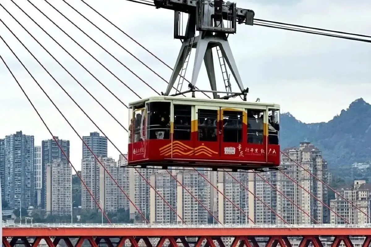 Yangtze Cable Car cabin in its red 重庆 livery crossing above the Yangtze River, with the Chongqing skyline and mountains behind.