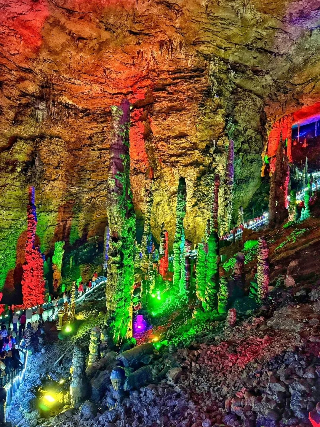 A large chamber in Yellow Dragon Cave, Zhangjiajie, filled with tall stalagmites lit in saturated red, green and orange floodlight, with visitors small on a walkway at the left.