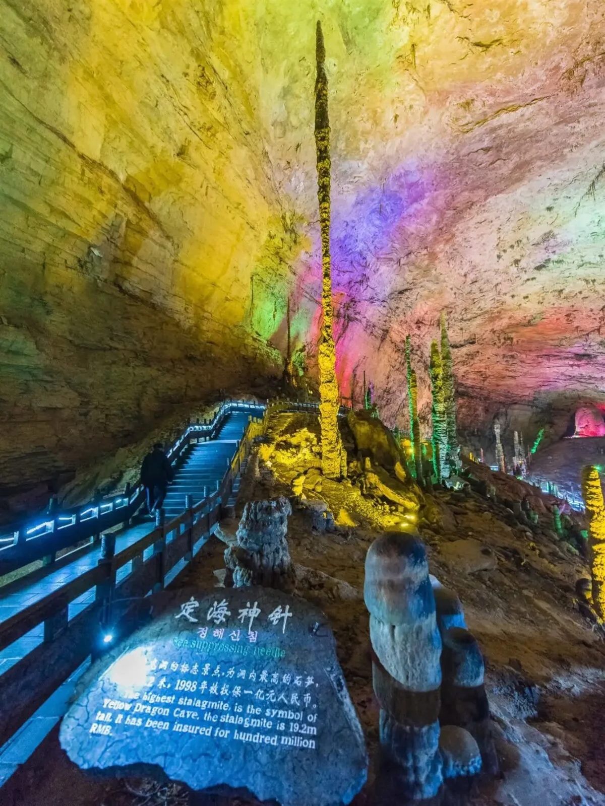 The Sea-Calming Needle stalagmite in Yellow Dragon Cave, Zhangjiajie — an extremely slender column running from floor to ceiling — with the site's interpretive stone in the foreground and a visitor climbing the stairway at the left.