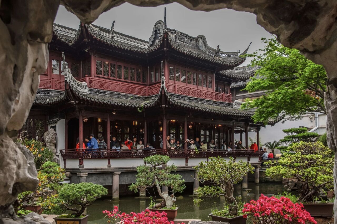 A pavilion in Yu Garden framed by rockery, bonsai and flowering azaleas, with visitors on the upper balcony.