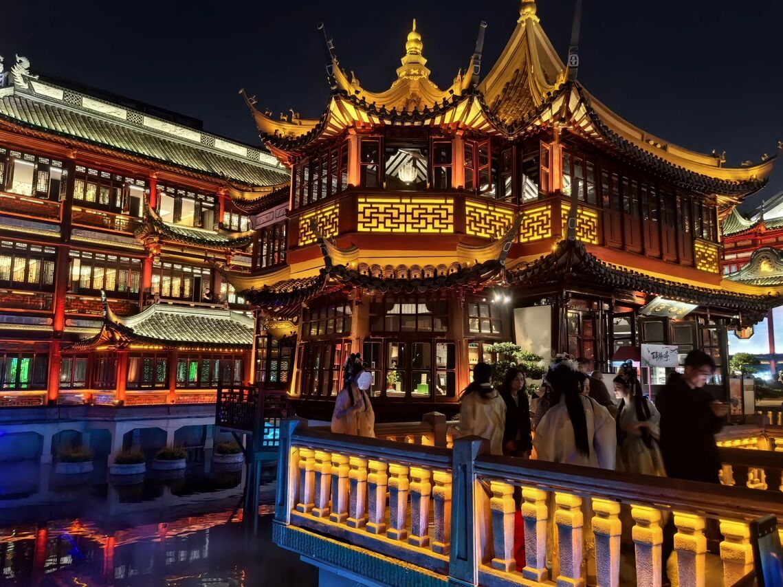 The Yuyuan Bazaar buildings lit gold against the night sky, with visitors crossing the illuminated bridge.