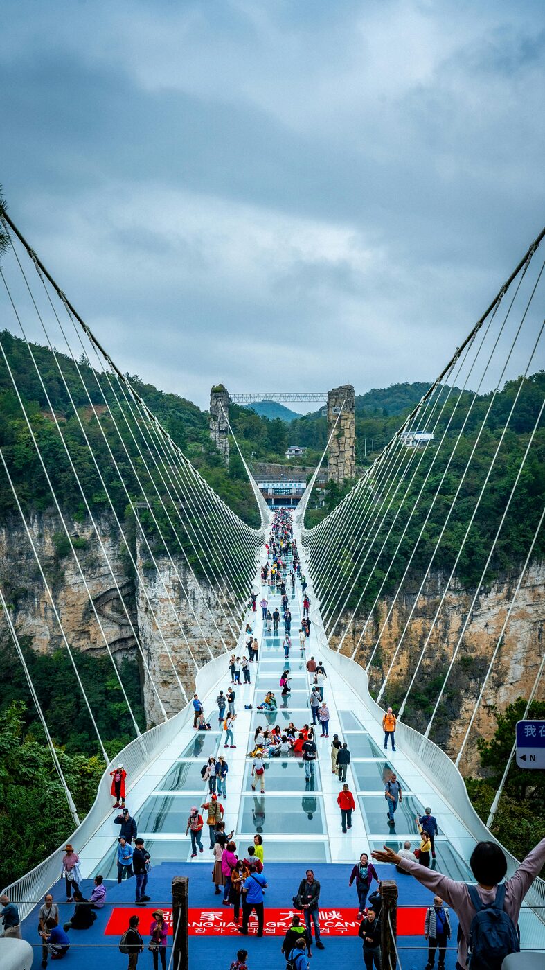 Looking along the deck of the Zhangjiajie Grand Canyon Glass Bridge — visitors crossing the transparent glass floor between the canyon walls.